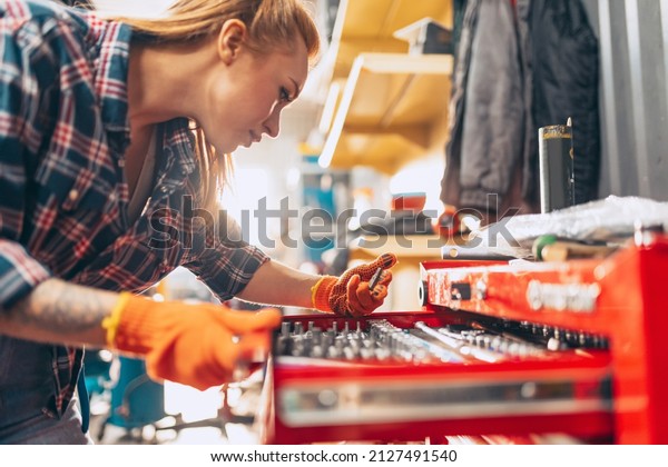 Car examining. Young red-headed girl, auto mechanic\
working at auto service station using different work tools. Gender\
equality. Work, occupation, car. Pretty girl in work clothes, plaid\
shirt