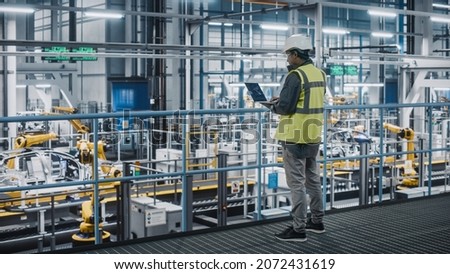 Car Factory Engineer in High Visibility Vest Using Laptop Computer. Automotive Industrial Manufacturing Facility Working on Vehicle Production with Robotic Arms Technology. Automated Assembly Plant.