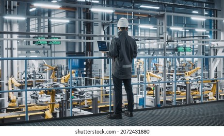 Car Factory Engineer in Work Uniform Using Laptop Computer. Automotive Industrial Manufacturing Facility Working on Vehicle Production with Robotic Arms Technology. Automated Assembly Plant.