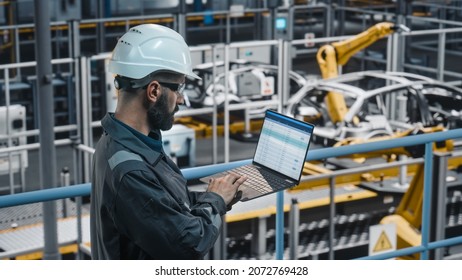 Car Factory Engineer In Work Uniform Using Laptop Computer With Spreadsheet Software. Working With Software At Automotive Industrial Manufacturing Facility Dedicated For Vehicle Production.