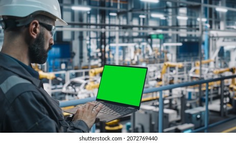 Car Factory Engineer In Work Uniform Using Laptop Computer With Green Screen Mockup Display. Working With Software At Automotive Industrial Manufacturing Facility Dedicated For Vehicle Production.