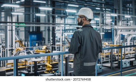 Car Factory Engineer In Work Uniform Using Tablet Computer. Automotive Industrial Manufacturing Facility Working On Vehicle Production With Robotic Arms Technology. Automated Assembly Plant.