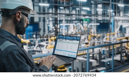 Car Factory Engineer in Work Uniform Using Laptop Computer with Spreadsheet Software. Working with Software at Automotive Industrial Manufacturing Facility Dedicated for Vehicle Production.