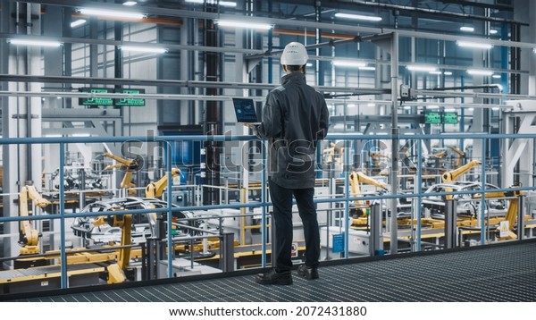 Car Factory Engineer in Work Uniform Using Laptop\
Computer. Automotive Industrial Manufacturing Facility Working on\
Vehicle Production with Robotic Arms Technology. Automated Assembly\
Plant.