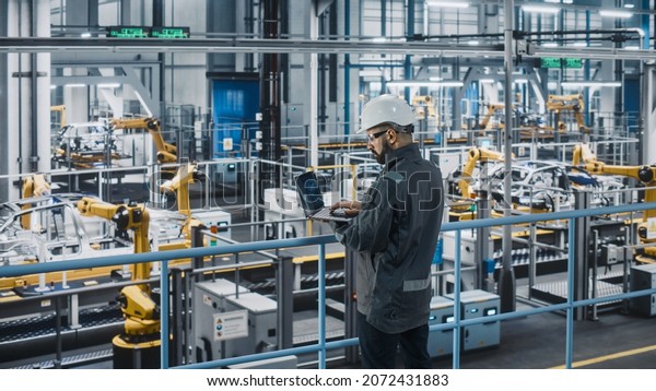 Car Factory Engineer in Work Uniform Using Laptop\
Computer. Automotive Industrial Manufacturing Facility Working on\
Vehicle Production with Robotic Arms Technology. Automated Assembly\
Plant.