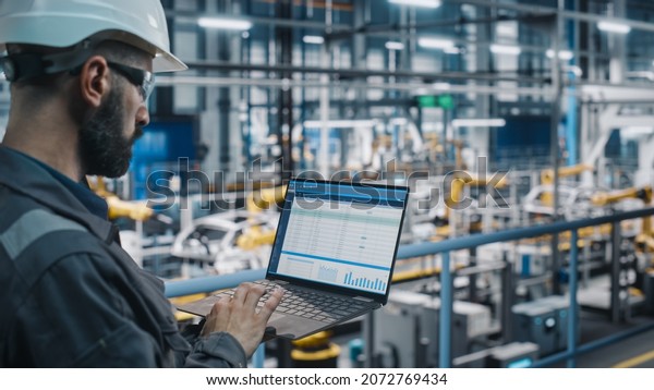 Car Factory Engineer in Work Uniform Using\
Laptop Computer with Spreadsheet Software. Working with Software at\
Automotive Industrial Manufacturing Facility Dedicated for Vehicle\
Production.