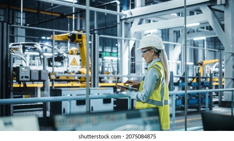 Car Factory: Female Automotive Engineer Wearing Hard Hat, Standing, Using Laptop. Monitoring, Control, Equipment Production. Automated Robot Arm Assembly Line Manufacturing Electric Vehicles. - Shutterstock ID 2075084875