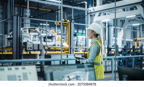 Car Factory: Female Automotive Engineer Wearing Hard Hat, Standing, Using Laptop. Monitoring, Control, Equipment Production. Automated Robot Arm Assembly Line Manufacturing Electric Vehicles. - Shutterstock ID 2075084878