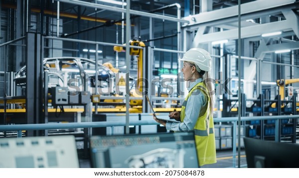 Car Factory: Female Automotive Engineer Wearing\
Hard Hat, Standing, Using Laptop. Monitoring, Control, Equipment\
Production. Automated Robot Arm Assembly Line Manufacturing\
Electric Vehicles.