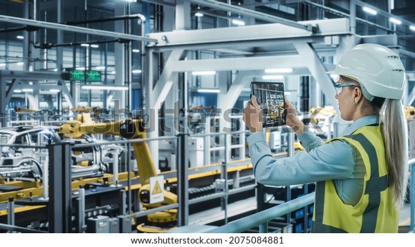 Car Factory: Female Automotive Engineer Using\
Augmented Reality Tablet Computer. Digitalization Scan, Monitoring\
Equipment Production. Automated Robot Arm Assembly Line\
Manufacturing Electric\
Vehicles