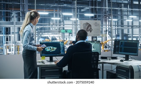 Car Factory: Female Supervisor Talks With Male Automotive Engineer, Using Computer To Design Advanced 3D Models For High-Tech Engines. Automated Robot Arm Assembly Line Manufacturing Electric Vehicles