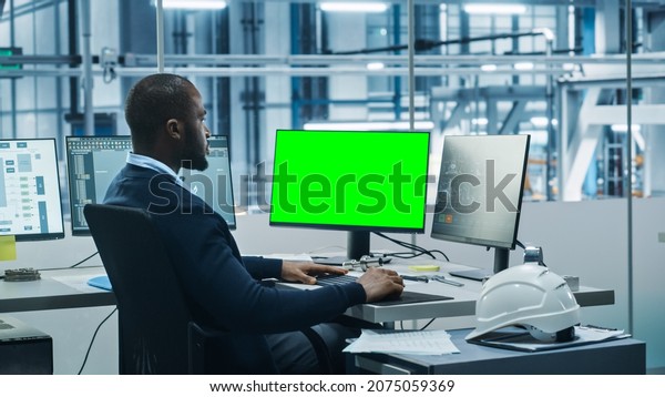 Car Factory Office: Black Engineer Works on a\
Computer, Two Monitor Screens Show Chroma Key Green Screen and\
Monitoring of Automated Robot Arm Assembly Line Manufacturing\
High-Tech Electric Vehicles