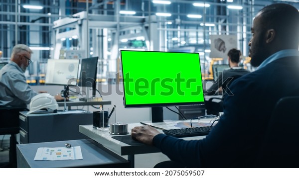Car\
Factory Office: Black Male Chief Automotive Engineer Sitting at His\
Desk Working on Green Screen Chroma Key Computer. Automated Robot\
Arm Assembly Line Manufacturing. Over Shoulder\
Shot