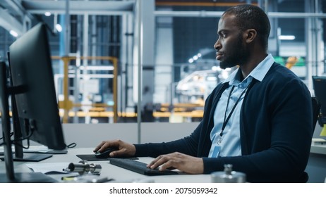 Car Factory Office: Portrait Of Confident Black Male Chief Engineer Working On Desktop Computer. Professional Technician In Automated Robot Arm Assembly Line Manufacturing High-Tech Electric Vehicles