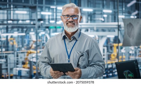 Car Factory Office: Portrait Of Senior White Male Chief Engineer Using Tablet Computer In Automated Robot Arm Assembly Line Manufacturing High-Tech Electric Vehicles. Looking At Camera And Smiling