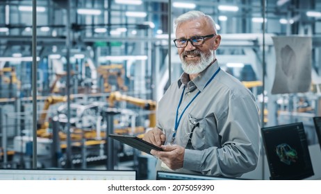 Car Factory Office: Portrait Of Senior White Male Chief Engineer Using Tablet Computer In Automated Robot Arm Assembly Line Manufacturing High-Tech Electric Vehicles. Looking At Camera And Smiling