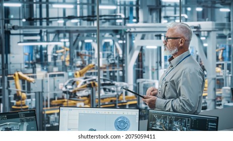 Car Factory Office: Portrait Of Senior White Male Chief Engineer Using Tablet Computer In Automated Robot Arm Assembly Line Manufacturing High-Tech Electric Vehicles. Side View Shot