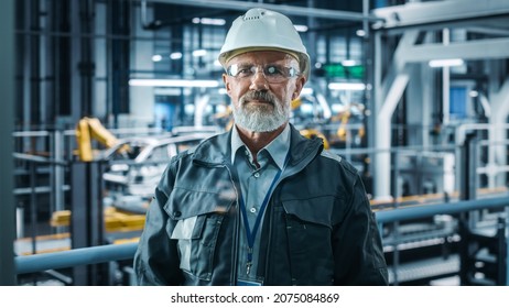 Car Factory Office: Portrait Of Senior White Male Chief Engineer Looking At Camera And Smiling. Professional Technician In Automated Robot Arm Assembly Line Manufacturing In High-Tech Facility