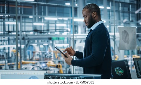 Car Factory Office: Portrait Of Successful Black Male Chief Engineer Using Tablet Computer In Automated Robot Arm Assembly Line Manufacturing High-Tech Electric Vehicles. Medium Side View