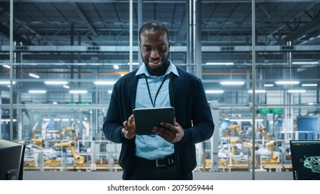 Car Factory Office: Portrait Of Successful Black Male Chief Engineer Using Tablet Computer In Automated Robot Arm Assembly Line Manufacturing High-Tech Electric Vehicles. Medium Shot