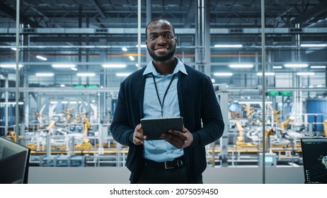 Car Factory Office: Portrait Of Successful Black Male Chief Engineer Using Tablet Computer In Automated Robot Arm Assembly Line Manufacturing High-Tech Electric Vehicles. Medium Looking At Camera