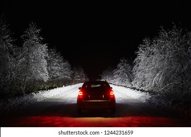 The Car Goes On A Snowy Road On A Dark Black Night In Winter. Headlights Brightly Illuminate The Path And The Forest After A Snowfall