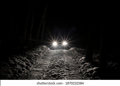 The Car Goes On A Snowy Road On A Dark Black Night In Winter. Headlights Brightly Illuminate The Path And The Forest After A Snowfall