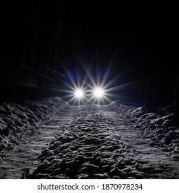 The Car Goes On A Snowy Road On A Dark Black Night In Winter. Headlights Brightly Illuminate The Path And The Forest After A Snowfall