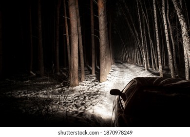 The Car Goes On A Snowy Road On A Dark Black Night In Winter. Headlights Brightly Illuminate The Path And The Forest After A Snowfall