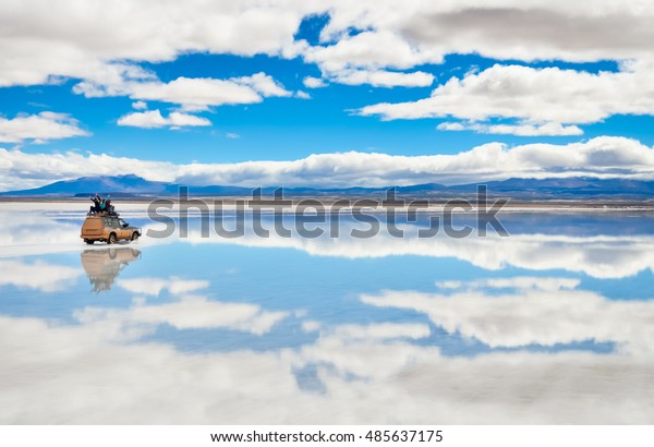 Car with happy people\
driving on the mirror surface of Salar de Uyuni in Bolivia with\
clouds reflection