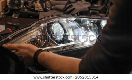 A car headlight in the hands of a man in close-up on a desktop background. An auto mechanic holds the headlight of the car. The concept of car repair in a car service. Dismantled headlight close-up.