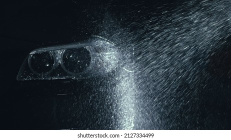 Car headlight wash. Washing modern vehicle body by high pressure jet wash hose water. Auto glass headlamp, angel eyes in drops. Close up of the spotlight of a black automobile as it gets cleaned.