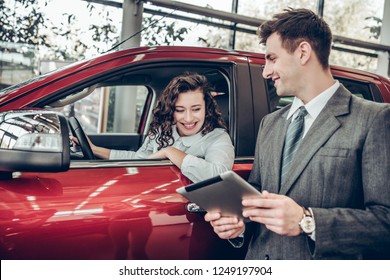 Car Info. Happy Young Woman Sitting In A New Car At Car Salon Professional Salesman Showing Them Information On Digital Tablet Couple Buying A Car Service Help Communication Clients Business Concept