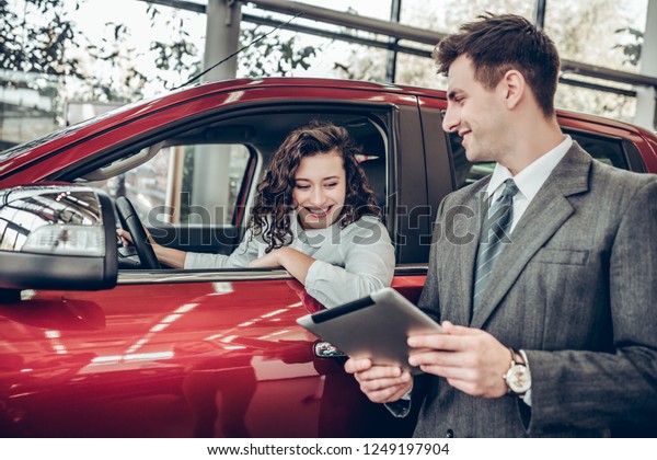 Car info. Happy young woman sitting in a new car\
at car salon professional salesman showing them information on\
digital tablet couple buying a car service help communication\
clients business concept