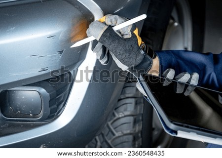 Car insurance claim process. Male hand with in protective gloves of car insurance agent or auto mechanic man using digital tablet and pen checking car scratch abrasion on front bumper