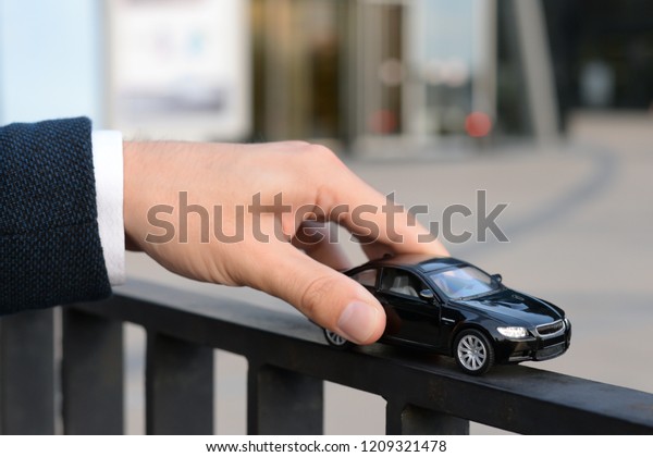 Car insurance. Car sale. Man in suit holding\
black car with protective gest. Black toy car in the men\'s hand on\
the city background.