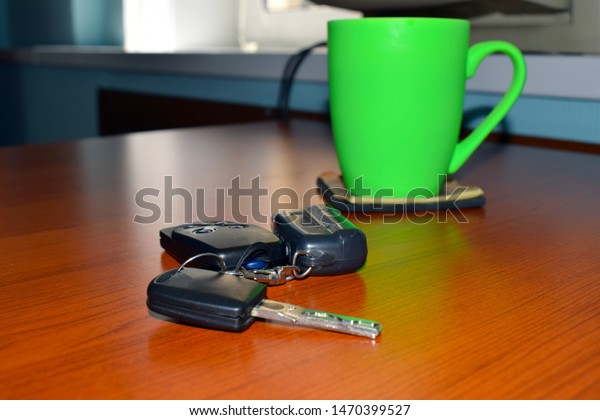 Car keys and key fob with alarm on a wooden office\
table and a bright green tea glass on a coaster. Black modern car\
door opener and keyless entry device with a car key and alarm fob\
on a wooden desk.