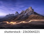 Car light trails on mountain road and high rocks at night in autumn in Passo Giau, Dolomites, Italy. Landscape with blurred light trails, hills, mountain peaks, purple sky with stars in fall at sunset