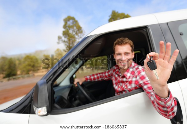 Car man driving rental car showing car keys\
happy looking at camera. Male driver on road trip in beautiful\
landscape nature. Focus on car\
key.