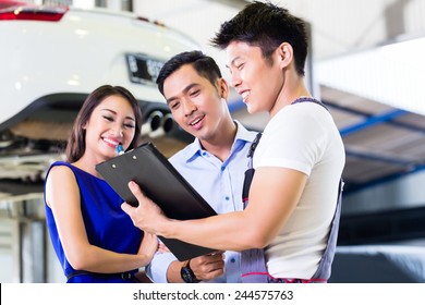 Car Mechanic And Asian Customer Couple Going Through Checklist With Auto On Hoist In The Background Of Workshop