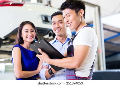 Car Mechanic And Asian Customer Couple Going Through Checklist With Auto On Hoist In The Background Of Workshop