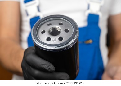 Car mechanic in blue jumpsuit holds oil filter of car in his hands and rotates it in black gloves. . Concept of oil change in engine in car, auto parts. - Shutterstock ID 2233331915