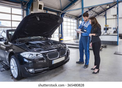 Car mechanic and customer stand next to the serviced car and looking through the checklist. The car had an annual checkup and stands in the garage with the hood open.