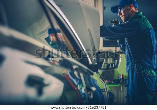 Car Mechanic Examining\
Vehicle Under the Hood. Caucasian Car Mechanic with Pro Flashlight\
at Work