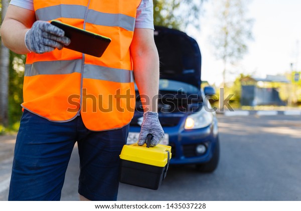 Car mechanic holding a box of tools and stands\
in front of car. Suitcase with tools and instruments for diagnosis\
in hands of car mechanic on background of a broken car. Mobile\
roadside assistance