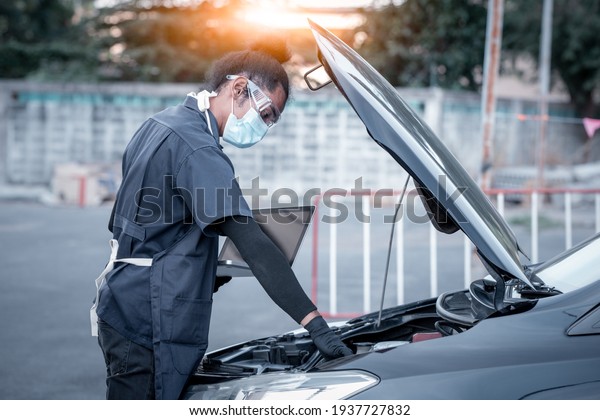 Car mechanic open car\
hood repairs and check problem system by laptop on hand to\
maintenance and fixed in garage .he wearing face mask to protect\
for virus and pollution.
