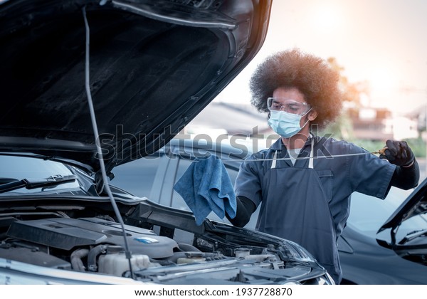 Car mechanic open car\
hood repairs system checking oil motor level for refill and clean\
in car garage service .he wearing face mask to protect from virus\
and pollution.