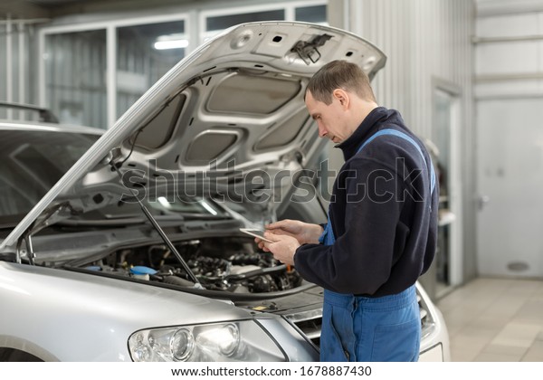 Car\
mechanic performing car diagnostics in the service center standing\
in front of the open hood with a diagnostic\
tablet