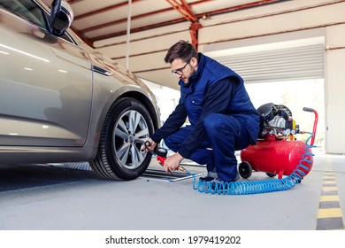 A car mechanic at the service station tries to inflate the tires on the car with the help of a compressor. A man in a blue work suit crouched next to a car in a workshop and works around car tires