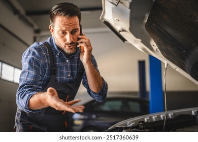 Car mechanic talk on the mobile phone, gesturing towards an engine in a garage setting, highlighting communication, repair work, and professional skills in automotive context - Powered by Shutterstock